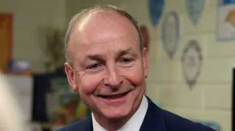 Michael Martin with short light hair, smiling at someone - he is wearing a navy suit with a white shirt. He is standing in a classroom with art on the walls. 