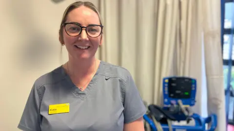 A woman with blonde hair, tied back, wearing glasses, a grey top and a yellow name card which says "Katie", smiles as she stands in a room at a hospital.