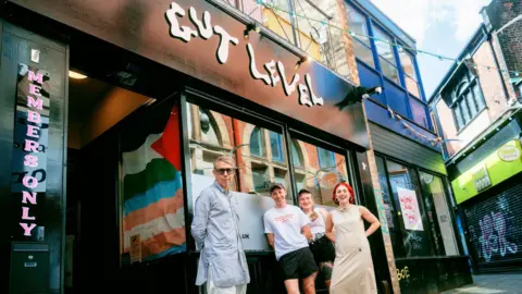 Republic Media The front of Gut Level music venue in Sheffield. The venue's name is above the entrance in a wavy font. Four people are stood outside it, looking at the camera.
