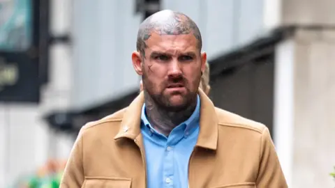 Tony Bartlett walks towards Bristol Crown Court. He is a large man with a shaved head and several tattoos on his neck and head, but not on his face itself. He is wearing a light blue shirt under a light brown jacket.