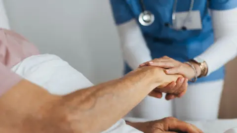 A medical professional wearing white trousers, a white long-sleeved top and blue scrubs holding an elderly person's hand. 