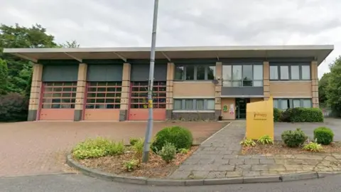 A two-storey fire station with three large doors for fire engines on the left hand side.