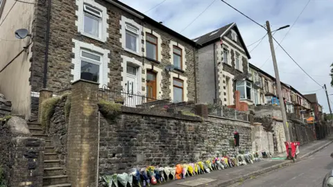 Google street view images of Graig Road. it is a row of brick row houses on one side, raised from the pavement to a small front garden. Bunches of flowers have been laid against the wall next to the pavement.