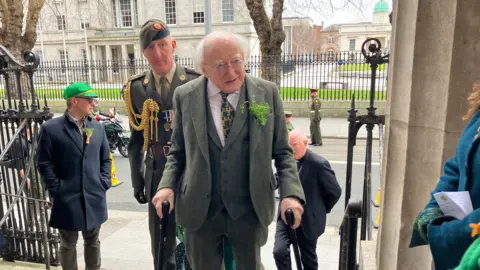 Irish President Michael D Higgins arrives for St Patrick's Day Mass in Dublin's Pro-Cathedral.  He has white hair and glasses and is wearing a grey three-piece suit with a sprig of shamrock pinned to his lapel.  He is walking with the aid of two walking sticks. An Irish soldier in green ceremonial uniform is walking up the steps behind Higgins. 