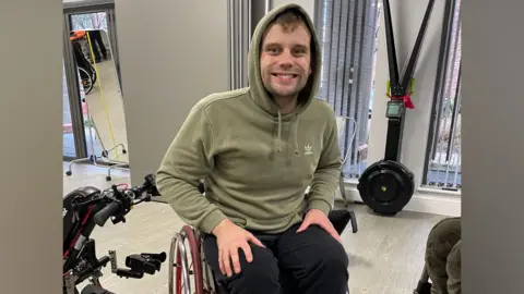 Liam Coffill Liam sits in his wheelchair at the gym and smiles at the camera. There are gym machines around him. He wears a sage green hooded top with the hood up.