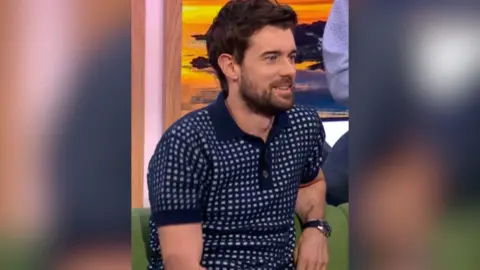 A man in a checked collared T-shirt sits on a green sofa