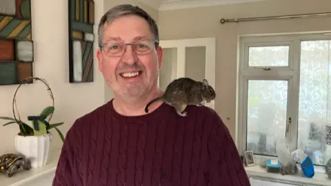 A man in glasses, wearing a burgundy jumper, has a rodent on his shoulder. The man is smiling at the camera