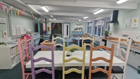 Clandeboye Primary A nurture room at the primary school which is bright and filled with pastel colours. There is a dining table with chairs around it, plus a kitchen, a desk with a computer on it, posters on the wall, and banners hanging from the ceiling.