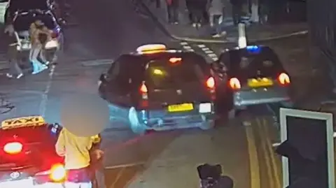 A car being driven half on a pavement and nearly hitting a taxi in a city centre at night. Pedestrians crossing the road are looking round.
