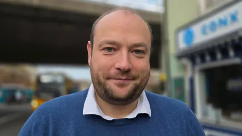 Qays Najm/BBC Martin Schmierer has a close-cropped beard and is wearing a blue jumper and open-necked shirt.