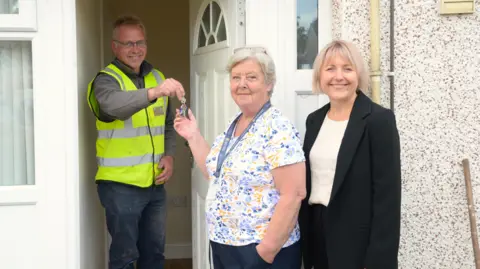 A man in a hi-vis jacket, opens the door to a council house. He is presenting keys to a woman wearing a floral blouse while another woman looks on, smiling.