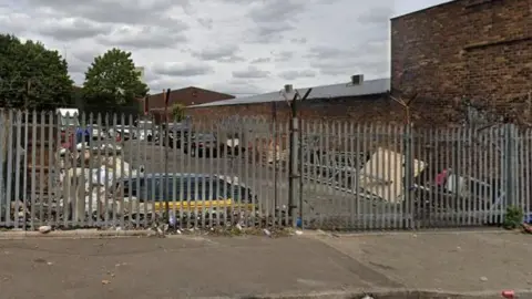 Google A cluttered car park pictured from outside the metal fence which surrounds it. Vehicle and rubbish can be seen around the car park. A large brick building is located to the right of the car park.