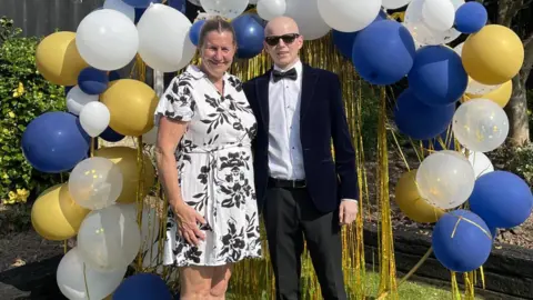 Family handout Owen with his mum Andrea. They are positioned in front of a decorative balloon arch made up of white, navy blue and gold balloons, along with some transparent balloons that contain gold confetti. Behind the balloon arrangement, there are gold metallic streamers hanging down.
Andrea is wearing a white dress with a black floral pattern and light-coloured sandals. Owen, on the right, is dressed formally in a dark suit jacket, white shirt, black bow tie, and black shoes. The background features green grass, a black wooden fence, and lush greenery including bushes and trees.