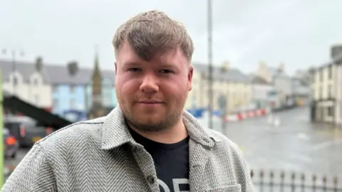 Ciaran Dallat is a young man with short, light coloured hair and a beard. He is wearing a grey and black patterned shirt with a black t-shirt underneath. He is stood in the town's Diamond, but the background is blurred.