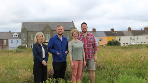 November Club  Four people stand in the wild grass in front of a row of coloured houses which make up the main street of the coastal village of Cambois.