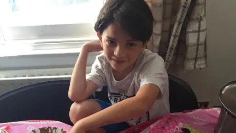 Oliver Archer Oliver Archer as a young boy. He is sat on a chair, wearing a grey t-shirt and blue shorts. He has brown hair. 