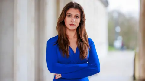 Cara Hunter, the SDLP MLA at Parliament Buildings at Stormont in Belfast. She is wearing a blue dress and has long dark hair. She is standing with her arms crossed and has a serious look on her face. 