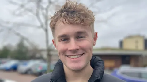 James is wearing a black puffer jacket. The background shows a parking lot with several cars, including a white car in the foreground and a blue car to the right.