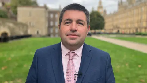 Shaun Davies is standing in front of Parliament buildings on a green lawn. He is wearing a navy blue suit jacket with light pink shirt and pink patterned tie.