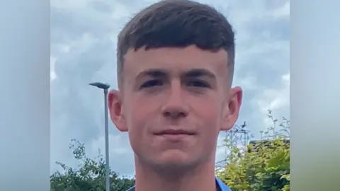 Submitted A young man looks at the camera with a slight smile. He has brown hair that falls over his forehead. A lamppost and hedges are in the background.