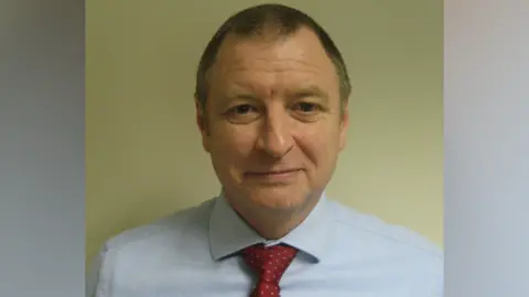 Disabled Motoring UK Graham Footer: A man who is standing against a cream wall looking directly at the camera. He is wearing a pale blue shirt and red tie.