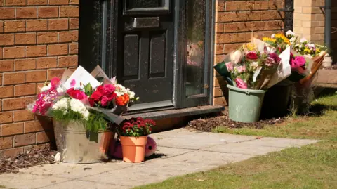 Martin Giles/BBC A large chrome bucket of flowers can be seen on a footpath to the left of a black door at the front of a property, with more flowers in pots on grass to the right.