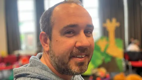 A man with brown/grey hair and brown/grey beard. He is sat looking into the camera over his right shoulder