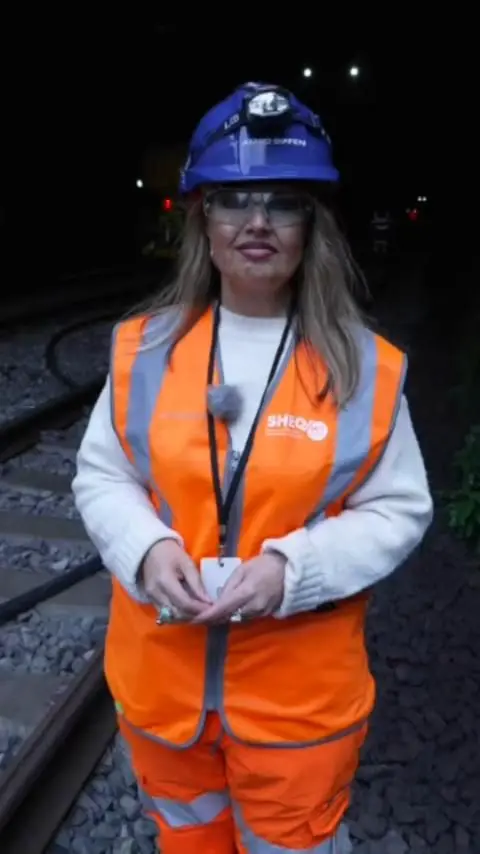 Rhiannon, wearing a blue hard hat, safety glasses and orange high viz wear, stands in front of a dark rail line.