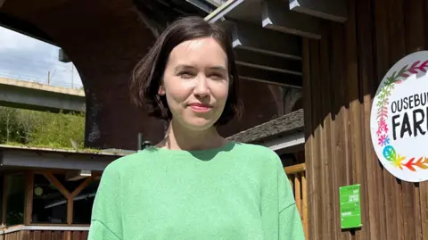 BBC/ANDY WATSON Katie Bates wearing a green jumper and blue denim jeans stands in front of Ouseburn Farm in Newcastle. The farm is situated undernead the Byker Bridge and the farm has the logo of the farm on the front. 