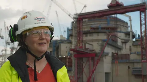 Nicola Fauvel is standing in front of the Hinkley plant that is under construction. She is wearing a red jumper, hi-vis jacket, safety goggles and a white hard hat with her name on. She also has earphones resting on the helmet. There are numerous cranes in the background.