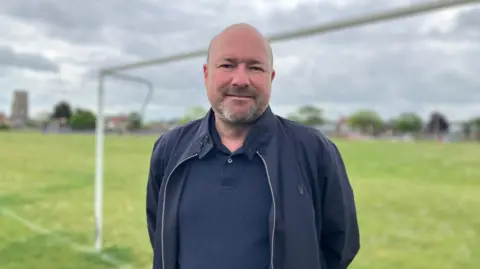 Owen Sennitt/BBC A man wearing a blue jacket and shirt smiles towards the camera in front of goal posts on a playing field
