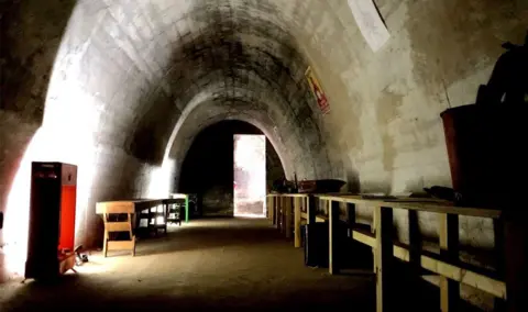 A public air raid shelter from World War Two. There are benches running the lenght of it and a stirrup pump can be seen in the foreground on one of them. Light is coming through a rectangular door of light is at the end. 