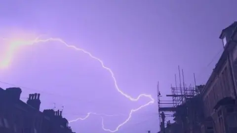 Cuppatea/Weather Watchers Lightning bolts in a purple sky above the silhouette of roofs and scaffolding.