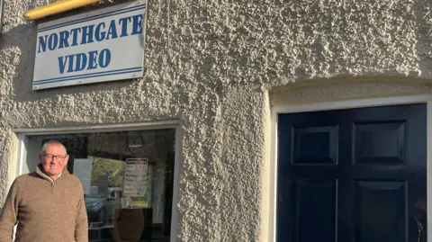 Rachael McMenemy/BBC Steve Webber stands outside the white building. It has a black door. He is smiling in the direction of the camera .There is a large window behind him, through which you can see DVDs. Above the window is a white sign with blue writing that reads 'Northgate Video'.