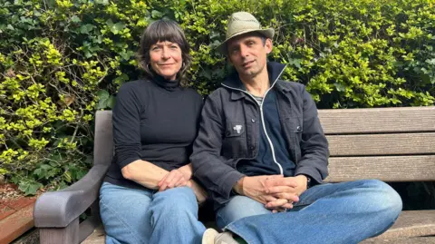 Kathryn Peters The image shows Mary Woodvine and Mark Jenkin sit together on a wooden bench in front of a thick green hedge. She has dark gair with a full fringe and is wearing a black top and blue jeans. Mark Jenkin has a dark blue jacket over a blue zip up top. He is wearing blue jeans and a Panama style hat.