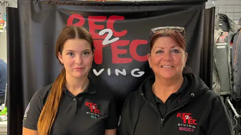Holly Nichols/BBC Two women, Mia Ellis-Kennedy and Maddy Kennedy, wearing black “Rec2Tec Diving” shirts stand side‑by‑side in a dive shop, smiling at the camera. A banner with the diving school’s logo hangs behind them, and shelves of equipment fill the background.