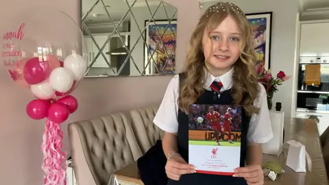 Girl with blonde hair hold a Liverpool Football Club poster. In the background is a kitchen. Pink and white balloons are to the left of her along with a mirror behind her.  