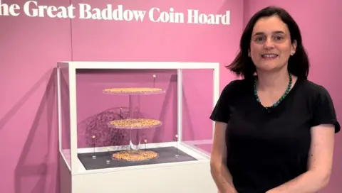 A woman wearing a black top and a dark beaded necklace with short hair. She is standing next to a small three tier stand with coins on each stand.
She is smiling at the camera.