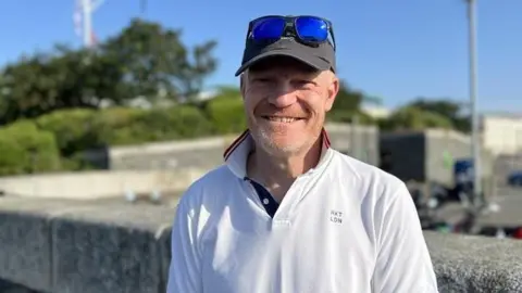 Man with ginger beard in baseball cap and glasses on his head, wearing a white polo shirt with collar popped up.