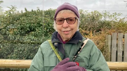 A woman sitting down in a bus shelter who is wearing dark sunglasses, matching purple hat and gloves and a light green coat with a red poppy.  