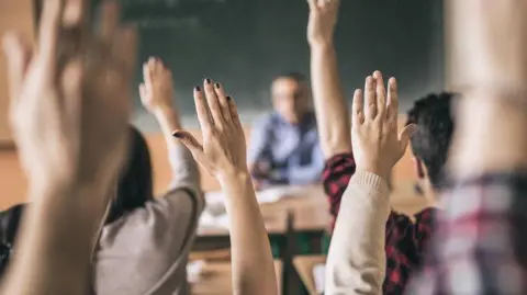 A general image of a classroom taken from behind students witting down at desks with their hands in the air. The background, which is blurred, shows a teacher sitting at a desk with a blackboard behind him.