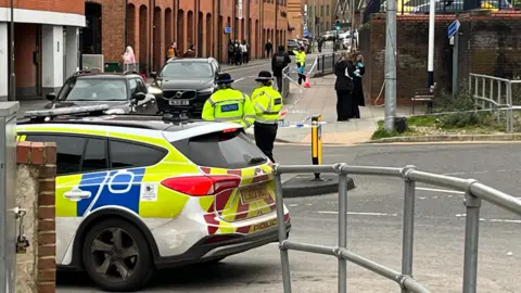 Two police officers wearing hi-vis jackets stand near a cordon with tape up, in a street. A police car is parked in front of the cordon. There is a red building and black cars behind the officers
