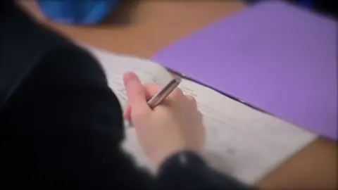 A young person holds a pen and writes on a white piece of paper or notebook