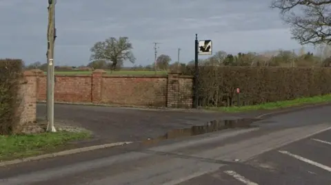 The entrance to what appears to be a farm drive. There is a hedge alongside and a road with white road marking in front. There is also a sign post and a telegraph pole on either side of the entrance.