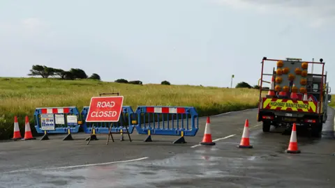 A road with barriers blocking it, and a lorry parked beside it. A sign in front of the barriers reads "road closed".
