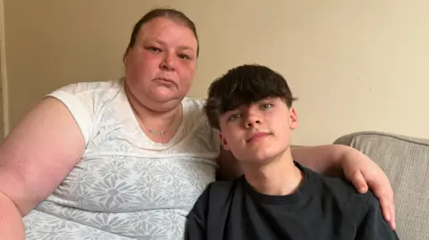 BBC/Jim Scott Julie Ann Fairless, left, and is wearing a white top with a flower pattern. She is sitting on a grey coloured sofa next to her son, Charlie, who she has her arm round. Charlie is wearing a black shirt and is smiling.