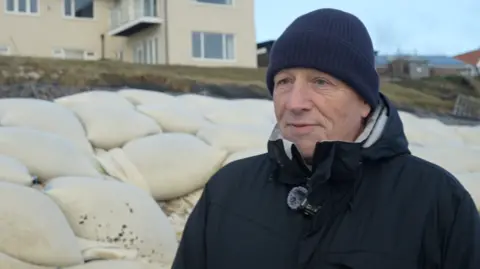 Tim Crisp looks to the left during an interview. There are sandbags piled up behind him. He is wearing a navy beanie hat and a black coat. A home stands on a cliff behind him.