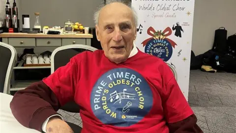 An elderly man who is mostly bald but with white hair at the back of his head, sitting at a table, which he is leaning on with his right arm. He is wearing a red shirt with a blue circle on with the words Prime Timers, World's Oldest Choir.