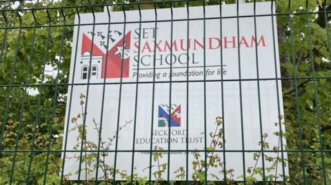Sign reading "Saxmundham School - providing a foundation for life" in front of a bush and covered by wire fence