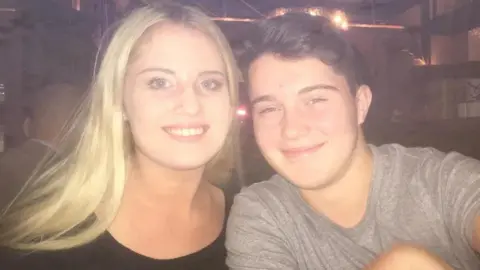 White family A young man with short, side-parted black hair wearing a grey t-shirt and a young woman in a black top sit next to each other in a dimly lit bar, smiling. The photo looks like it was taken some time ago, and there is a slight haze to the image.
